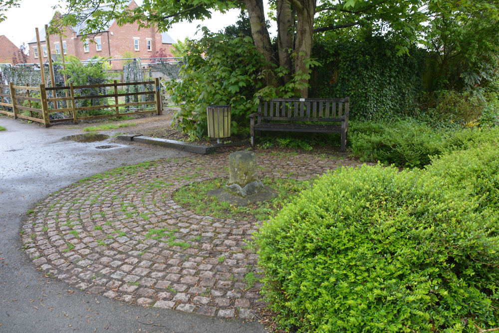 At first sight, it's easy to miss the stump of this old village cross, as the area in which it sits is busy with benches, bollards (at the entrance to the small park behind) and telegraph poles, etc. The remains of the cross shaft sit in the middle of a circular paved area, with the stump secured in place with concrete and lead flashing.