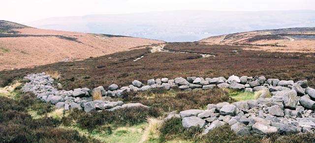 Bronze Age enclosure of rubble walling. The site was excavated in the mid 1980's and the wall and hut circles were partially reconstructed. Prior to this the wall was only to be found by searching in the heather.