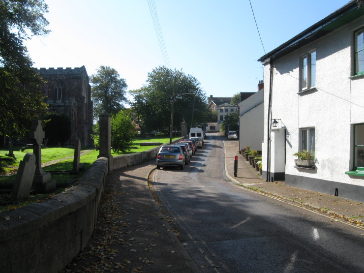 A view taken opposite the lane to access the well; looking back at the cottage and the top of the road, with the parish church on the left here.