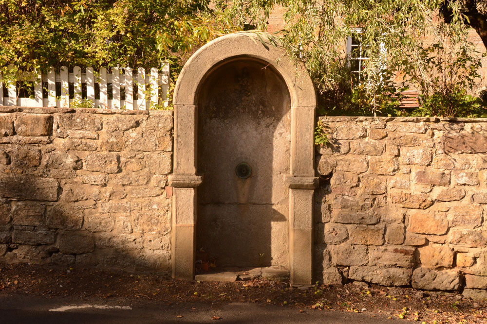 The well/fountain housing is built into the wall of the Old Post Office, at the side of the B6343.