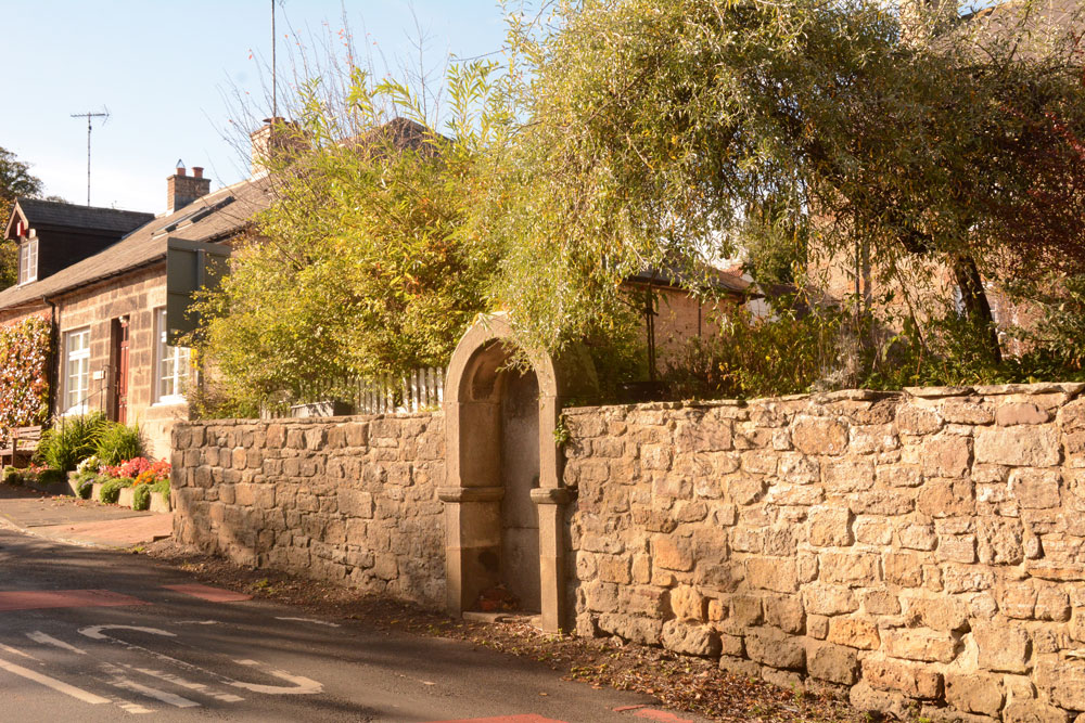 Photo showing the general location of the well/fountain housing, approaching from Morpeth. It is built into the wall of the Old Post Office, just before the Old Blacksmith's Cottage and the junction of the B6343 with Font Side.