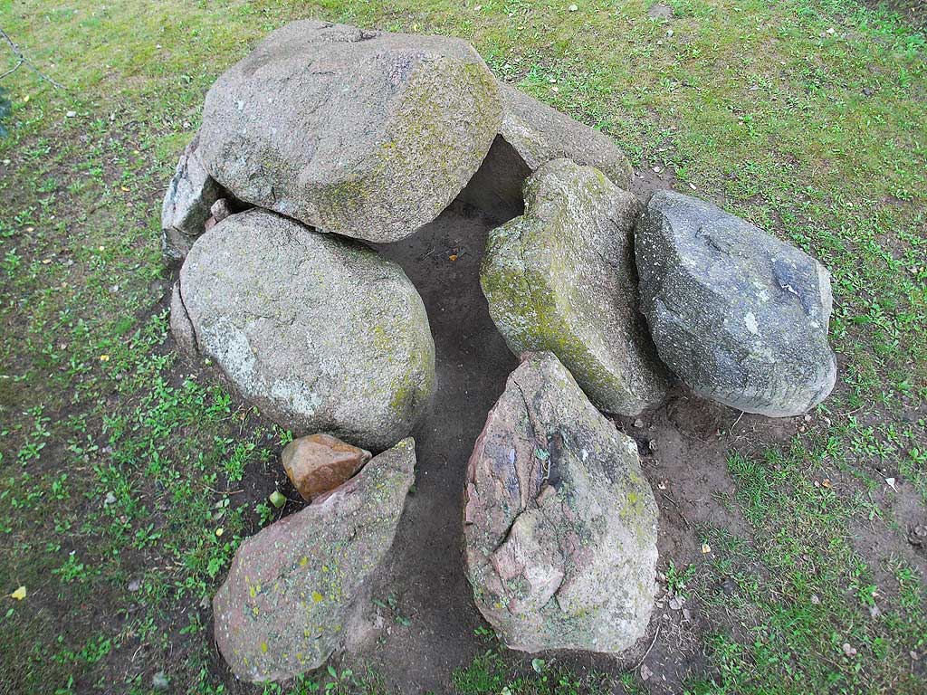 Dolmen in NE-SW orientation, with entrance at the west corner.
The chamber is set with four uprights, two bigger ones on the long sides.
In the west corner is a gap, two stones of the passage are also in situ.
One capstone, 1.8m x 1.5m x 0.8m, is resting on the chamber;
another, smaller one leans outside against the chamber.

(View from pole)