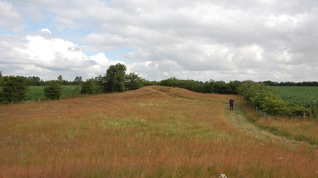 Round Barrow, 3 m x 17 m. From the east side leads an 8 m long passage (uncovered in 1931) into a chamber.
The chamber is 3 m in E-W, 1.50 m br., 1.40 to 1.60 m high.
Chamber is set by six carrying stones, two in the south side,
one in the west end, the north side two long stone, which the west has shifted somewhat and pushed a portion inward, especially at the top. 
The entrance from S-E is 0