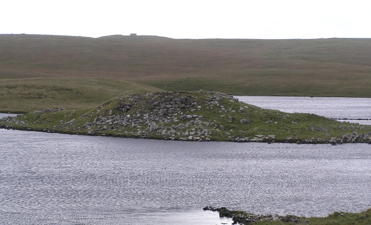View south over the Loch of Houlland and its broch.

The next broch south in the chain around the Shetland coast was sited on the hill at Sae Breck which can be located on this picture from the square remains on the horizon of the WWll building which was built on the same site.  There are no readily identifiable remnants of the Sae Breck broch, but its position can be located through the trig po
