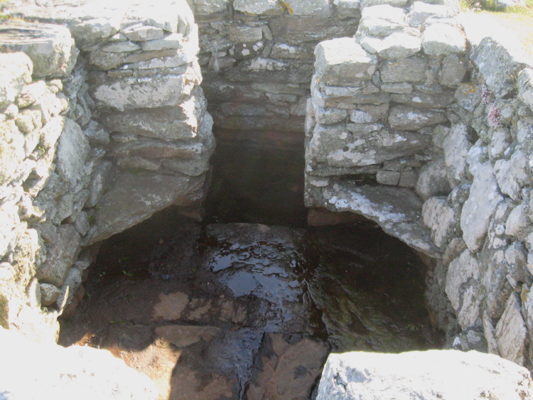 Standing on the steps, looking down into the chamber of the well, showing the ledges at each corner for seats and the small pool through the gap at the west of the area.